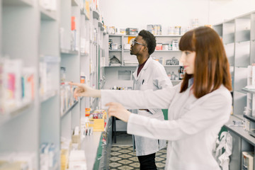 Fototapeta premium Portrait of a young Caucasian female pharmacist, male African colleague working with drugs in the background. Focus on man