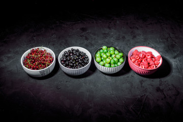 A four bowls overflowing with summer berries like currants, raspberries, blueberries and gooseberries on dark background