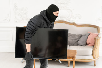 Thief with black balaclava stealing modern expensive television. Luxury apartment with stucco. Man...