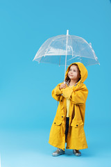 Happy funny child with transparent umbrella posing on blue studio background. Girl is wearing...