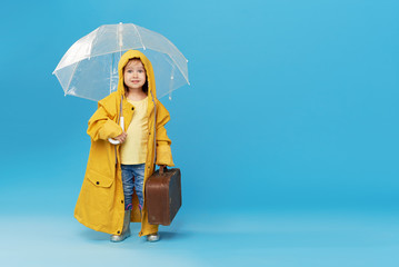 Happy funny child with transparent umbrella posing on blue studio background. Girl is wearing...
