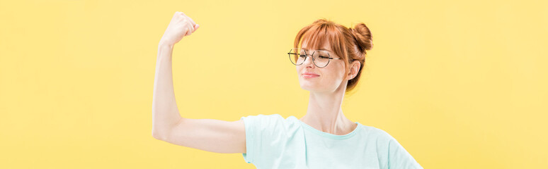 panoramic shot of confident pleased redhead girl in glasses and t-shirt holding fist up isolated on yellow