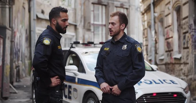 Two handsome friendly policemen talking near the police car together patroling the city at daytime. Police on duty.