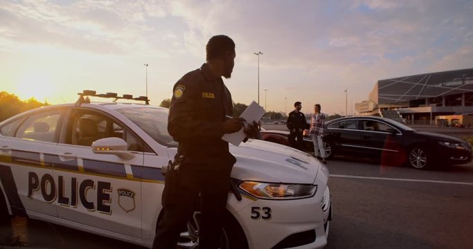 Young man black policeman leaning on car hood writing a fine doing his job under sunset in the city. On background offended people talking with the cop to resolve an issue.