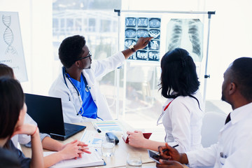 Afro American doctor shows and explains MRI results to his professional team. Group of international doctors or interns have a meeting and making notes at hospital room. Health care concept.