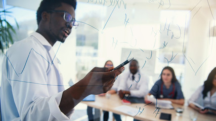 African professor at medical conference in modern clinic teaches his students. Doctor writing on board some formulas for interns in conference room at hospital at sunrise.