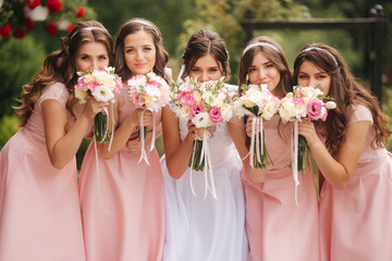Fototapeta premium Happy bride with bridesmaid hold bouquets and have fun outside. Beautiful bridesmaid in same dresses stand by the charming bride in long wedding dress