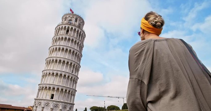 Back of delighted young woman overlooking magnificent Leaning Tower of Pisa. Low angle view of stylish girl tourist walking in Piazza dei Miracoli. Traveling to Tuscany, Italy