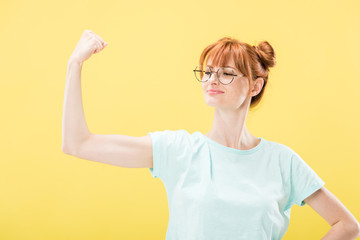 confident pleased redhead girl in glasses and t-shirt holding fist up isolated on yellow
