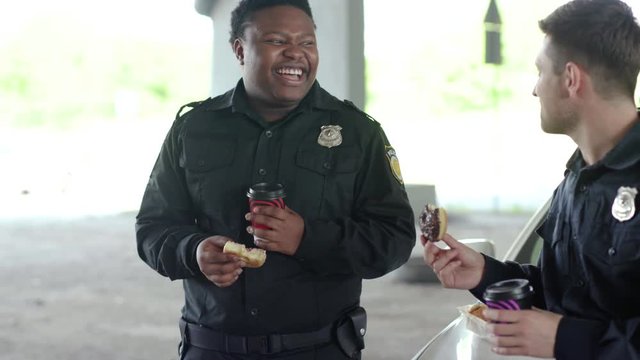 Face cute smiling two policemen drinking coffee and eating donut with appetite talk near patrol car help happy african american break rest smilegangster uniform funny teamwork portrait slow motion
