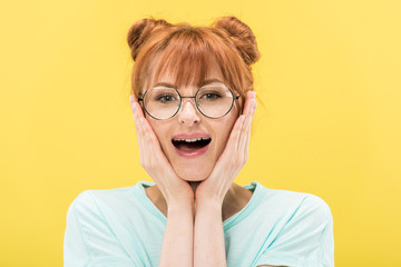 front view of excited redhead girl in glasses touching face and looking at camera isolated on yellow