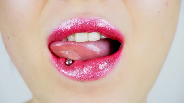 Extreme close-up of the lips of a young woman. A woman sexually licks bright full lips. She has a tongue piercing.