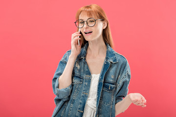 attractive redhead girl in glasses and denim jacket talking on smartphone isolated on pink