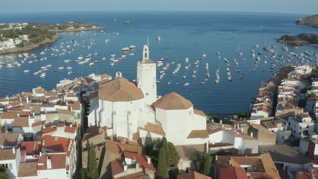 Drone flight around church of St. Mary in coastal town of Cadaques on background of beautiful blue bay with anchored yachts. Aerial view of old historic building and houses along coastline