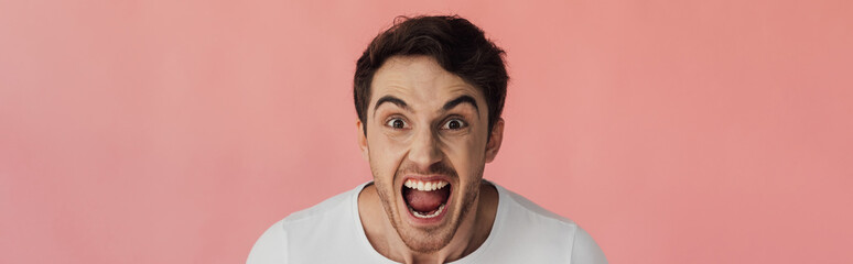 panoramic shot of angry young man looking at camera and screaming isolated on pink