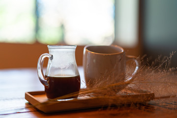 A tray with cup of hot drink and a glass jar is on a wooden surface in front of the window