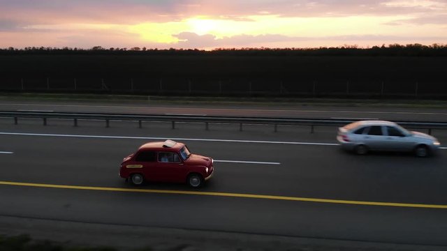Great aerial footage of red small retro car drive on 4 lane motorway in sunset or sunrise with turned headlights. Side close up shot