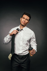 african american businessman with cash in pocket holding coffee to go and untying tie on dark background