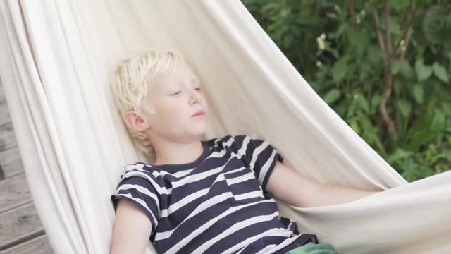 Happy blond boy lies in a hammock and sings songs in the summer on a farm