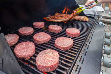 man's hand with tungs over an outdoor Propane gas grill on a deck with hamburgers and rack of pork ribs and flames
