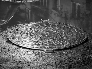 Metal sewage cover in the flooded city, reflecting buildings in the dirty water in black and white.