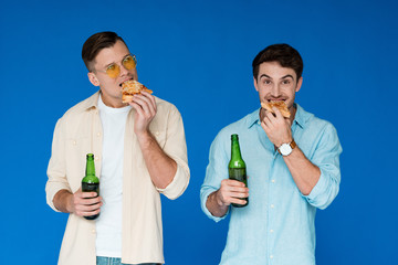 front view of two friends holding bottles of beer and eating pizza isolated on blue