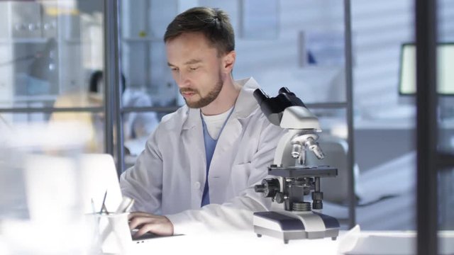 Panning waist-up shot of Caucasian technician busy at work in hospital lab, analyzing tissue specimen under microscope and filling in data on laptop