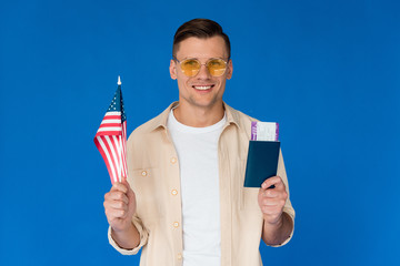 front view of smiling tourist in sunglasses holding passport with air ticket and american flag isolated on blue