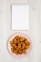 Fototapeta na wymiar Overhead view, homemade fried plantains on a pink plate, blank notepad on a white wooden background. Flat lay, top view, from above. Copy space.