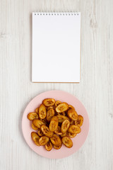 Overhead view, homemade fried plantains on a pink plate, blank notepad on a white wooden background. Flat lay, top view, from above. Copy space.