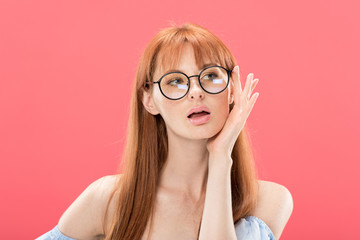 curious redhead woman looking at camera and touching glasses isolated on pink