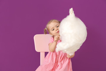 Cute little girl with cotton candy sitting on chair against color background