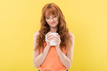 smiling redhead girl holding cup of coffee isolated on yellow