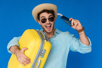 excited traveler in safari hat and sunglasses holding yellow suitcase and passport with air ticket isolated on blue