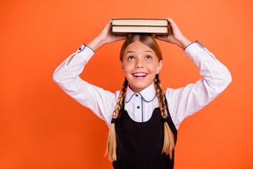 Close-up portrait of her she nice attractive charming cute cheerful cheery playful pre-teen girl holding on head book first September grade isolated over bright vivid shine orange background