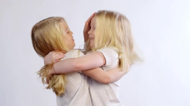 Waist-up shot of vivacious 6-year-old blonde twin girls hugging while posing in studio on white background, smiling for camera and then turning to each other