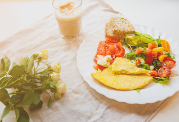Healthy breakfast. Omelette with tomatoes and rucola salad. Salmon with wholemeal bread. Cappuccino and roses on the rustic linen background. Ideal morning flat lay.