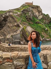 Young girl in blue dress with long hair and earrings rises on the island Gaztelugatxe on the coast of the Bay of Biscay, Spain, Basque Country