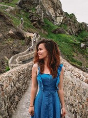 Young girl in blue dress with long hair and earrings rises on the island Gaztelugatxe on the coast of the Bay of Biscay, Spain, Basque Country