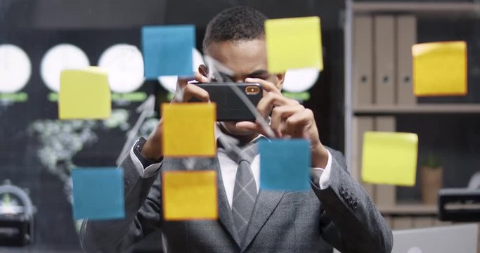 Young african man are standing in a modern office, look at the glass board with sticky notes and takes a picture on smartphone.