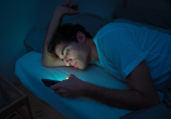 Young caucasian sleepless man bored in bed surfing on the Internet addicted to mobile phone