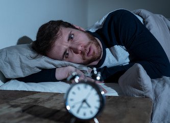 Desperate stressed young man whit insomnia lying in bed staring at alarm clock trying to sleep