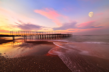 Fototapeta premium Opuszczona plaża o zachodzie słońca. ogromny księżyc nad wodą i stare żelazne molo.
