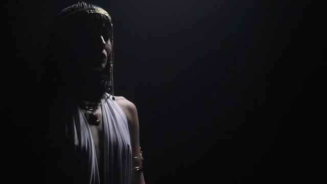 Cleopatra in white dress, with the crown is standing in low lighting