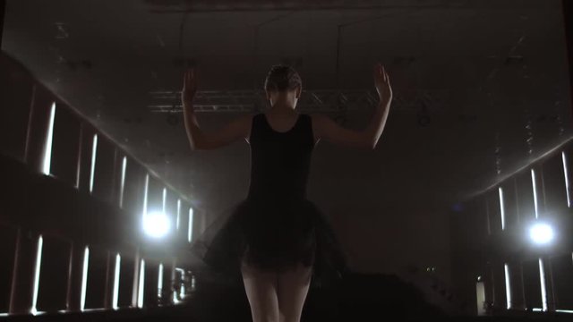 Close-up of ballet dancer as she practices exercises on dark stage or studio. Ballerina shows classic ballet pas. Slow motion. Flare, gimbal shot