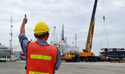 Rear view of engineer in reflective cloth with safety helmet is working and showing thumb up gesture on shipping port area at harbor 