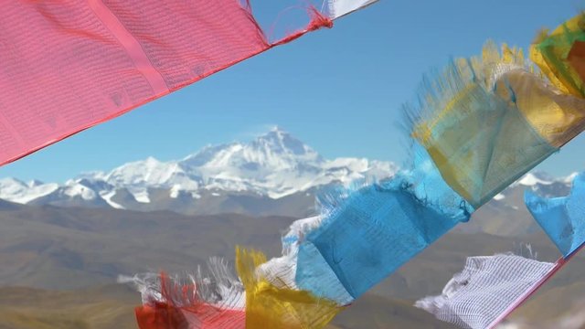 SLOW MOTION, CLOSE UP, DOF: Spectacular view of the snow covered Everest behind colorful prayer flags flapping in the wind. Mount Everest towering above the smaller barren hills in beautiful Tibet.