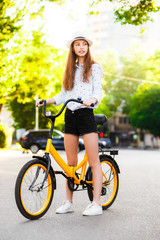 Girl teenager in a hat with a yellow bike walks through the summer city.