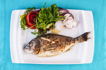 Mediterranean dish, European cuisine. whole fish baked in the oven, served with a salad of vegetables, greens, arugula, onion rings, tomato and lemon