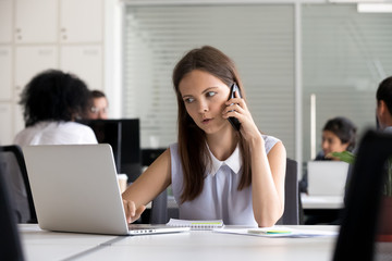 Young female intern talking on phone, consulting using laptop